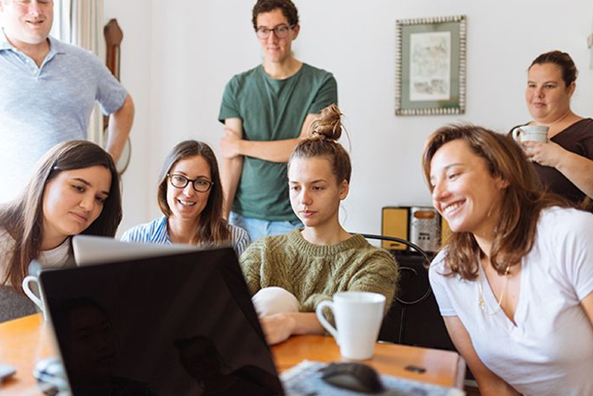 four women seated at a table in front of a computer while two men and another woman views their work standing behind them