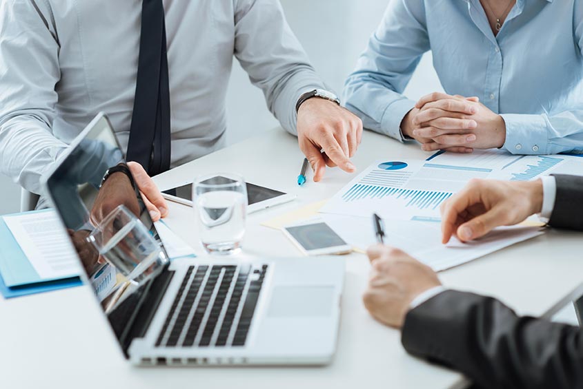 two men and a woman sitting a table looking over paper business documents and an open laptop