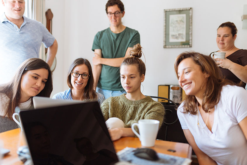 four women seated at a table in front of a computer while two men and another woman views their work standing behind them